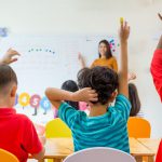 Preschool kid raise arm up to answer teacher question on whiteboard in classroom.