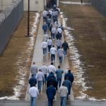 A line of prison inmates walk across the prison yard next to a barbed wire fence