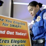 A brightly colored sign reads Are you eligible for SNAP? Find out today. In the background a woman in a TSA uniform stands with her head down