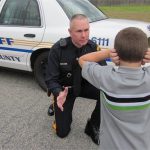 A police officer in uniform kneels in front of a young boy standing with his hands over his ears