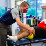 In a physical therapy clinic, a college student wearing a mask bends over and touches the knee of an older man sitting facing him.