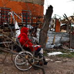 A woman in a red coat sits on her wheelchair beside houses destroyed by Russian shelling amid Russia's invasion of Ukraine, in Sumy