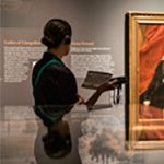 A person with dark hair pulled into a bun walks through the Museum of the American Revolution with a printed guide and a pair of headphones.