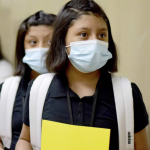 Medium-skinned pre-teen girls in school uniforms carrying backpacks wear masks.