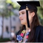A college-age woman with long dark hair wears a black graduation gown and mortar board and a flower necklace.