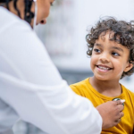 A boy with curly dark hair smiles as a healthcare provider in a white coat listens to his chest through a stethoscope.