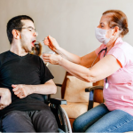A young adult with short dark hair and some facial hair, seated in a wheelchair, opens his mouth as an older woman in a face mask with a stethoscope feeds him with a spoon.