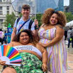 Chloe Leon, seated in a wheelchair, holds a fan with the many colors of many pride flags. A person in a dress with big, curly hair and a bright smile rests a hand on Leon’s arm. A person with short hair wearing a tie-dyed t-shirt with a Keith Haring-style illustration stands behind Leon’s wheelchair.