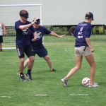 John Weaver and another player take defensive stances on a soccer field. They are both wearing goggles that block their vision. A third person dribbles a soccer ball toward them.