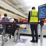 A person in a wheelchair waits with a staff member in a safety vest in the disabled section of the baggage carousel.