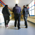 Three people walk down a school hallway, seen from behind. The person in the middle wears a jacket with the word Security on the back.