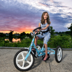 Alyssa Wolfe straddles an adaptive tricycle along the Milford river walk, which is a paved surface. Behind her the sun sets with bold colors against clouds. She has long dark hair and long, thin legs.