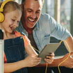 A smiling man in a chambray shirt leans over the shoulder of a girl wearing yellow headphones plugged into a tablet.