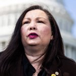 U.S. Senator Tammy Duckworth stands before the U.S. Capitol dome. She has straight dark hair and wears red lipstick and a black blazer.