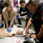 Healthcare workers receive training on CPR. Right is an instructor wearing a uniform. Center, an Asian person with short hair wearing blue nitrile gloves leans over from a chair to observe. A person with longer blond hair pulled up and away leans over the CPR dummy while wearing protective goggles and blue nitrile gloves.