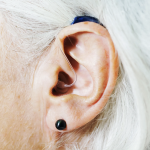 Close up of a hearing aid in a person's ear. They have smooth white hair that tucks behind the ear and their skin indicates that they are an older person. They wear on small black globe earring.