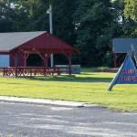Camp Lenape sign in Felton with red-painted wood pavilion with picnic tables in the background. The sign has red letters on a pyramidal blue wooden structure.