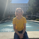 Riley Brooker sits on the edge of a fountain. Sunlight glows above over a building and backlights Brooker.. Brooker has feathery shoulder-length mid-tone hair and wears a yellow t-shirt, jeans and glasses.