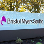 A large exterior sign for Bristol Myers Squibb sits in a landscaped area under large trees.