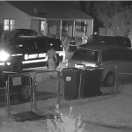 Black and white still from nighttime video footage shows two New Castle County Police SUVs with headlights on in a residential street. The grainy footage looks over a small front yard with chain link fence, trash cans left for pickup and a few parked vehicles. A few police officers and another person are visible.
