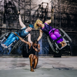 From left, Laurel Lawson, Jerron Herman and Alice Sheppard, rehearsing Kinetic Light’s “Wired." Lawon has short blue hair and wears a shiny blue costume, while strapped into a wheelchair and suspended by a strap. Similarly, Sheppard has curly blond hair and wears a purple costume. Herman, kneeling between the two, wears a black top and coppery tights that are almost indistinguishable from his skin.