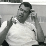 Carl Croneberg in the 1960s. Shown in grainy black and white, he wears a white button down shirt, has a buzz cut and signs with one hand by his temple and the pinky of his right hand up while the other fingers are down.