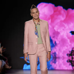 A masculine presenting person with short hair molded into a faux hawk wears a dusky pink suit, grey shirt and colorful silk neck scarf while walking down a catwalk runway.