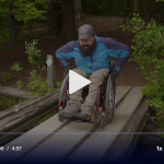 A person with a beard in a wheelchair rolls across a bridge made of board with gaps that could easily trap the wheelchair's wheels.