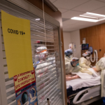 A hospital room with a COVID-19 sign posted on it. Healthcare workers gather around a patient inside.
