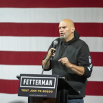 John Fetterman wearing a Carhartt hoodie holds a microphone at a podium in front of a giant U.S. flag.