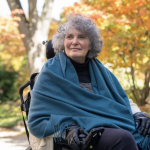 Lisa Iezzoni has short curly grey hair and uses a wheelchair. She wears a scarf and gloves. The trees behind her show their fall colors.