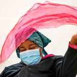 A woman with medium skin tone wearing a turquoise kerchief and a face mask waves a pink scarf in the air.