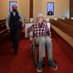 A person in a wheelchair rolls through a church with red carpeting and wooden pews.