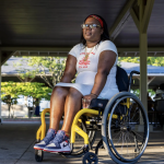 A young adult woman in a yellow wheelchair sits on a covered concrete pad.