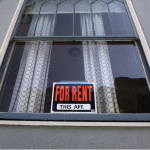 Leaded glass window in a grey stucco building with a For Rent sign posted.