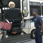 A Paratransit driver helps a passenger in a wheelchair into a bus using a lift.