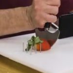 A hand working a crescent shaped rocking cutting tool to cut a strawberry, which is held in place by metal spikes on a adaptive cutting board.