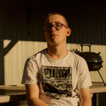 A young adult with light-toned skin and very short hair sits on a sunny porch. They wear a Puma t-shirt and glasses.