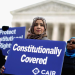 A person wearing a hijab holds a sign that says "Constitutionally Covered" in front of a neoclassical building with columns and pediment