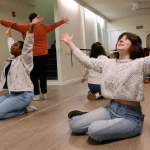People in a dance studio sit on the floor or stand and spread their arms wide