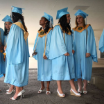 High school students stand in groups, wearing baby blue mortar boards and gowns, some with yellow regalia hoods.