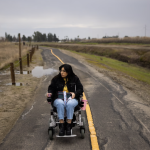 A young adult with long dark hair and glasses sits in a wheelchair along a desolate country road. Grey clouds fill the sky.