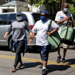 Three people in casual clothing wearing face masks walk across a street. One holds the arm of another, who is one of two carrying bags with supplies for their community outing.