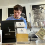 A young adult with short dark hair and goatee works a register, a tip jar in front.