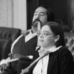 Black and white photo of Judy Heumann and Ed Roberts seated at a table with microphone.