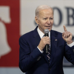 President Biden is an older person with short white hair, wearing a blue blazer and white button down. He holds a microphone and points up with his free hand.