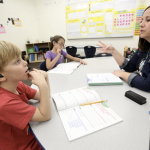 A teacher and young student sit across a classroom table from one another. The student has a hearing aie. The teacher holds three fingers out while speaking.