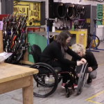 A person in a wheelchair gets assistance from a repair person in a bike shop.