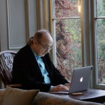 An older adult sits at a table by a large window while working on a laptop