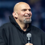 John Fetterman is a middle-aged man with a bald head and a short greying beard. He wears a black hoodie and holds a microphone.